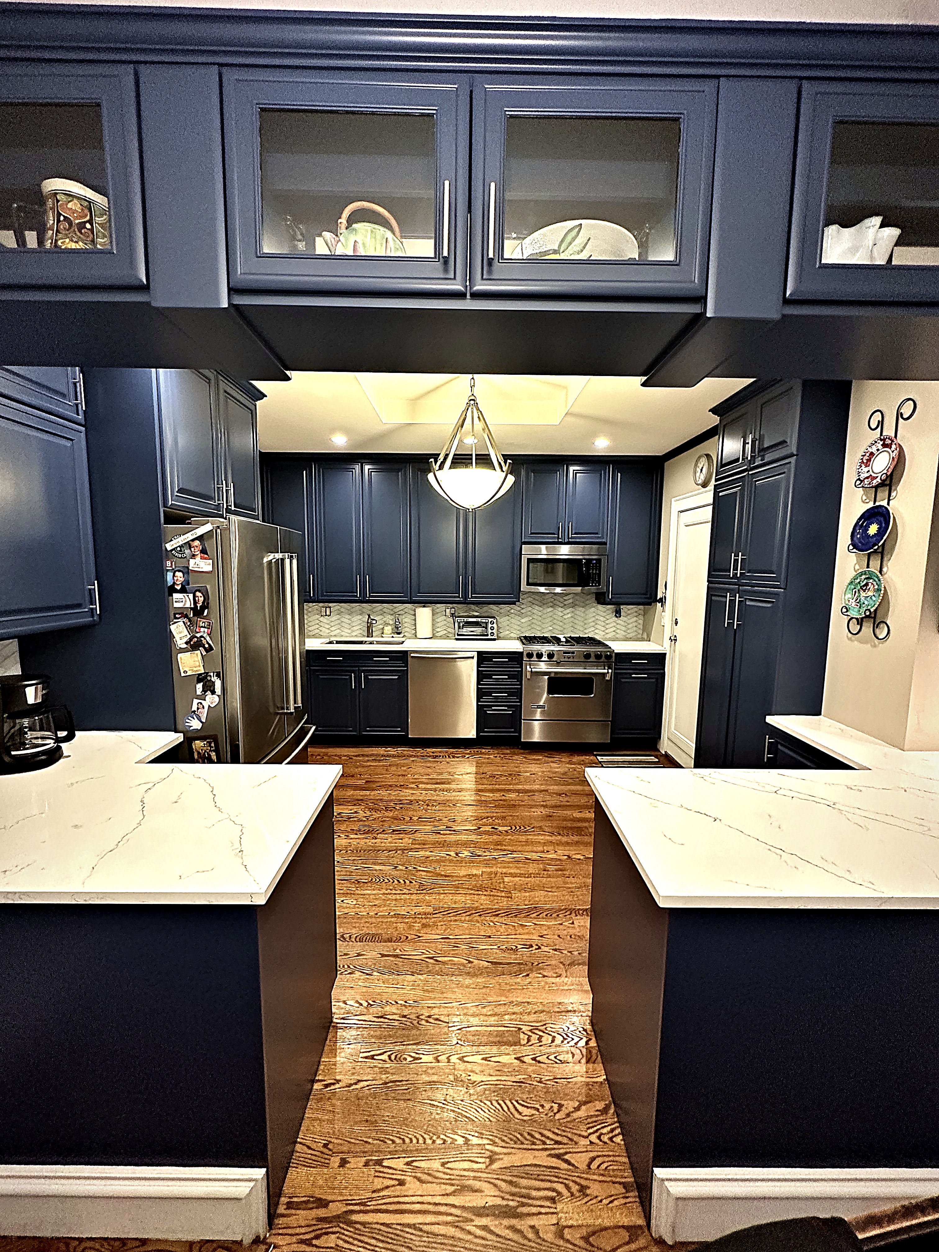 A kitchen with deep navy cabinets, glass upper doors, white stone countertops, and warm oak floors, refinished by GK Painting.